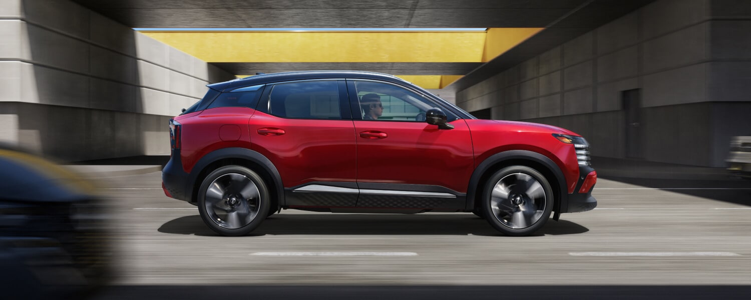 Side of a red 2026 Nissan Kicks SR driving down a road.