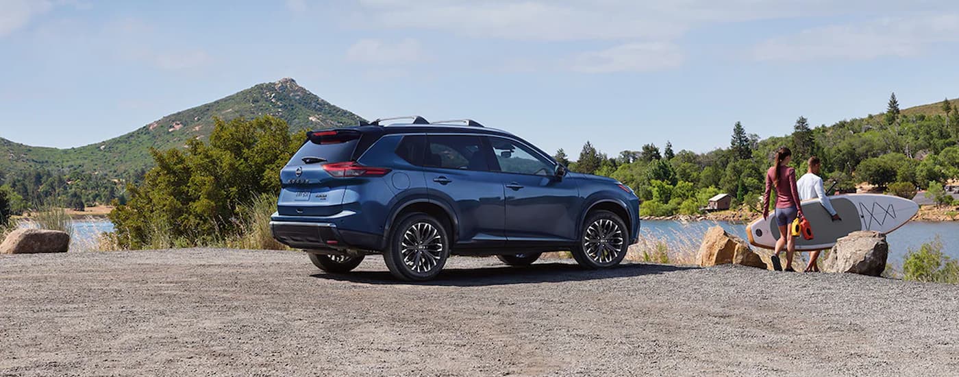A blue 2024 Nissan Rogue Platinum is parked on gravel near a lake.